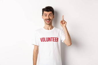 英俊的年轻的男人。志愿者t恤的想法提高手指建议指出站白色背景