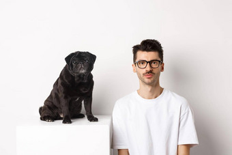 图像英俊的赶时髦的人眼镜坐着黑色的可爱的哈巴狗狗盯着相机白色背景