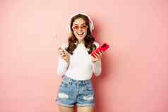 时尚的夏天女孩跳舞智能手机萝莉波普听音乐耳机站太阳镜粉红色的背景