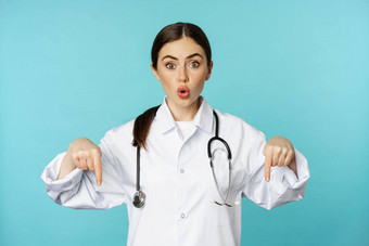 热情的医疗保健工人年轻的女人医生白色外套指出手指微笑显示药店广告医疗促销蓝色的背景