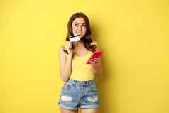 在线购物时尚的浅黑肤色的女人女人持有智能手机信贷卡支付应用程序移动电话应用程序购买smth站黄色的背景