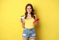 时尚的年轻的浅黑肤色的女人女人持有信贷卡智能手机支付购买订单商店站黄色的背景