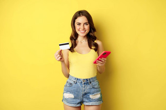 时尚的年轻的浅黑肤色的女人女人持有信贷卡智能<strong>手机支付</strong>购买订单商店站黄色的背景