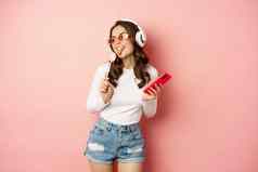 时尚的夏天女孩跳舞智能手机萝莉波普听音乐耳机站太阳镜粉红色的背景