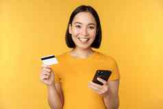 在线购物快乐的亚洲女孩持有信贷卡智能手机支付订单移动电话站黄色的背景