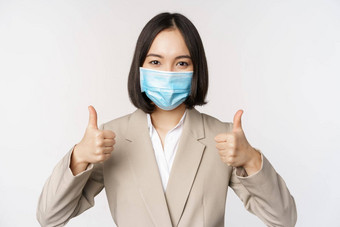 冠状病毒工作场所概念图像亚洲女售货员公司工人医疗面具显示拇指标志微笑很高兴白色背景