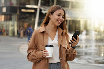 企业家年轻的忙女人智能手机应用程序办公室工人在户外卡布奇诺咖啡咖啡杯购物购物中心体系结构现代城市背景