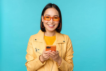 微笑亚洲女孩太阳镜智能<strong>手机</strong>应用程序持有移动电话站蓝色的背景