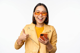 快乐时尚的朝鲜文女孩穿太阳镜显示拇指持有智能<strong>手机</strong>移动电话应用程序推荐应用程序在线商店白色背景