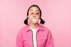 时尚的亚洲女孩吹泡泡糖泡沫咀嚼口香糖穿太阳镜摆姿势粉红色的背景