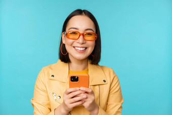 肖像热情的亚洲女人太阳镜移动电话微笑笑快乐持有智能<strong>手机</strong>站蓝色的背景