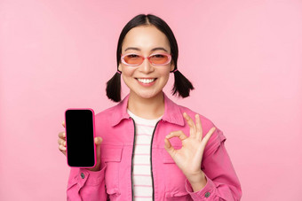 热情的年轻的亚洲女人显示标志微笑很高兴移动电话屏幕智能<strong>手机</strong>应用程序站粉红色的背景