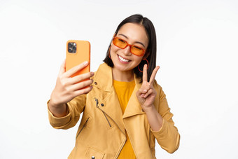 时尚的美丽的亚洲女孩太阳镜采取自拍智能手机摆姿势照片持有移动电话白色工作室背景