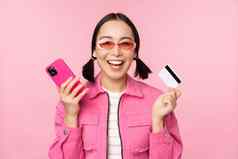 现代美丽的亚洲女孩笑微笑移动电话信贷卡购物在线支付智能手机站粉红色的背景