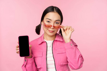 时尚的朝鲜文女孩年轻的女人太阳镜显示智能手机屏幕移动电话应用程序接口网站站粉红色的背景