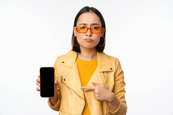 肖像伤心亚洲女人太阳镜指出手指移动电话应用程序接口显示智能手机应用程序站白色背景