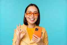 快乐朝鲜文女孩太阳镜显示标志持有移动电话智能手机应用程序推荐应用程序站蓝色的背景