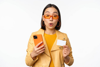 在线购物时尚的亚洲女模型太阳镜持有信贷卡移动电话微笑快乐站白色背景