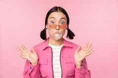 时尚的亚洲女孩吹泡泡糖泡沫咀嚼口香糖穿太阳镜摆姿势粉红色的背景