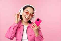 时尚的亚洲女孩跳舞智能手机听音乐耳机移动电话应用程序微笑笑摆姿势粉红色的背景