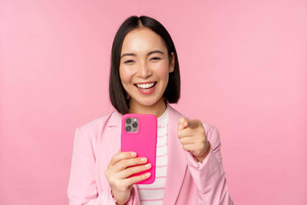 快乐亚洲女商人笑指出手指记录视频采取照片智能手机移动电话粉红色的背景
