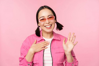 肖像友好的亚洲青少年女孩太阳镜波手微笑问候手势站粉红色的背景
