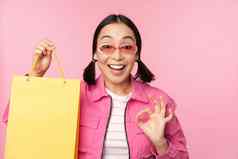 购物时尚的亚洲女孩太阳镜显示袋商店微笑推荐出售促销商店站粉红色的背景