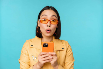 图像亚洲女孩太阳镜惊讶深刻的印象记录视频采取照片智能手机站蓝色的背景