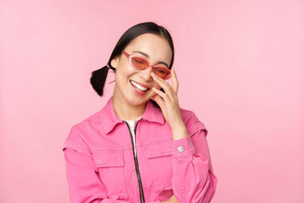 关闭肖像时尚的亚洲女人太阳镜微笑受宠若惊笑卖弄风情的站粉红色的背景
