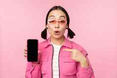 热情的亚洲女人时尚的衣服太阳镜指出手指移动电话屏幕显示智能手机应用程序站粉红色的背景