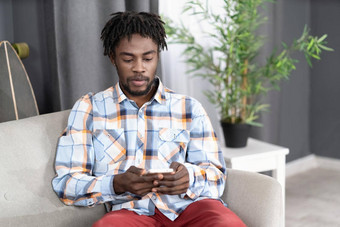 集中感兴趣非洲美国男人。智能手机检查社会媒体更新信息肖像年轻的男人。持有智能手机坐着沙发社会媒体概念