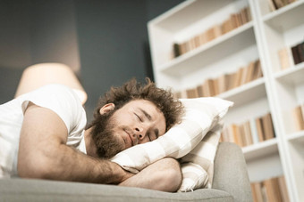 睡觉沙发英俊的有胡子的年轻的男人。拥抱枕头长小时工作晚些时候近生病的年轻的男人。睡觉沙发上采取药物距离工作概念