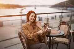 视频调用美丽的年轻的女人智能手机杯咖啡法国女人穿红色的贝雷帽坐着阳台餐厅咖啡馆背景秋天城市城市