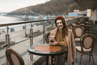 快乐年轻的女孩坐着在户外街餐厅阳台微笑持有咖啡杯子肖像时尚的年轻的女人穿秋天外套红色的贝雷帽在户外