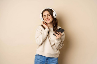 快乐高加索人女人听音乐耳机智能手机应用程序跳舞有趣的站米色背景