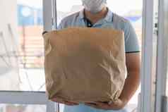 交付男人。穿脸面具蓝色的统一的处理袋食物给客户前面房子邮递员表达杂货店交付服务