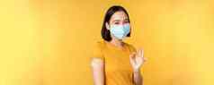 疫苗接种科维德健康概念快乐亚洲女孩显示穿医疗面具乐队援助肩膀冠状病毒疫苗拍摄黄色的背景