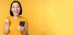 快乐的亚洲女人庆祝持有移动电话赢得实现目标智能手机站黄色的背景