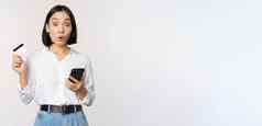在线购物概念图像惊讶亚洲女孩持有信贷卡智能手机惊讶难以置信相机白色背景