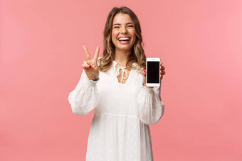 肖像卡哇伊乐观快乐年轻的女孩白色衣服显示移动电话显示和平标志笑感觉快乐的很高兴<strong>分享</strong>太棒了应用程序<strong>链接</strong>粉红色的背景