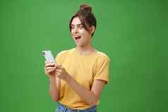 女人阅读令人惊讶的令人满意的消息智能手机开放口兴奋微笑惊讶惊讶手机屏幕摆姿势绿色背景休闲黄色的t恤