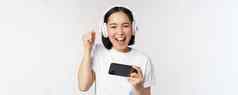 快乐亚洲女孩玩家玩移动电话看智能手机穿耳机站白色背景