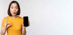 兴奋亚洲女孩显示移动电话屏幕标志推荐智能手机应用程序站黄色的T恤白色背景