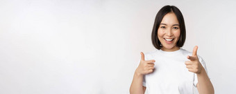 图像微笑亚洲女孩指出手指相机选择邀请祝贺站T恤白色背景