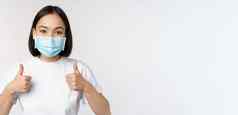 科维德健康人概念年轻的快乐朝鲜文女人医疗面具显示拇指站T恤白色背景