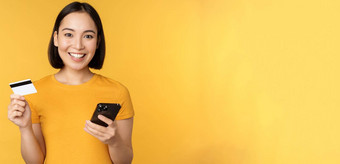 在线购物快乐的亚洲女孩持有信贷卡智能手机支付订单移动电话站黄色的背景