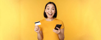 在线购物快乐的亚洲女孩持有信贷卡智能<strong>手机</strong>支付<strong>订单</strong>移动电话站黄色的背景