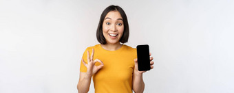 兴奋亚洲女孩显示移动电话屏幕<strong>标志推荐</strong>智能手机应用程序站黄色的T恤白色背景