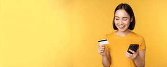 在线购物微笑亚洲女孩信贷卡移动电话应用程序支付非接触式订单智能手机应用程序站黄色的背景