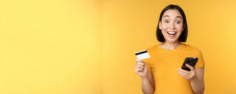在线购物快乐的亚洲女孩持有信贷卡智能<strong>手机支付</strong>订单移动电话站黄色的背景